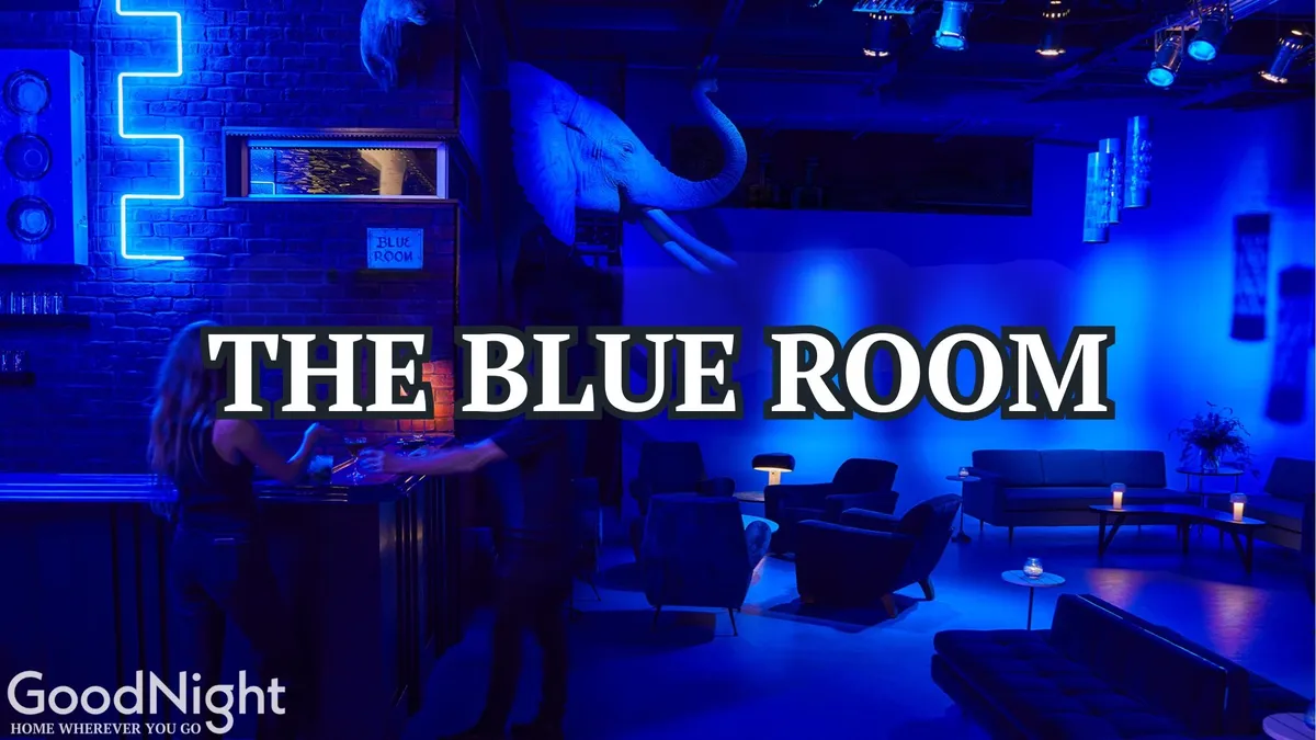 A moody, intimate interior featuring blue lighting, a sophisticated bar area, comfortable seating, and eye-catching wall art. The scene suggests relaxing with cocktails in a stylish, urban setting.