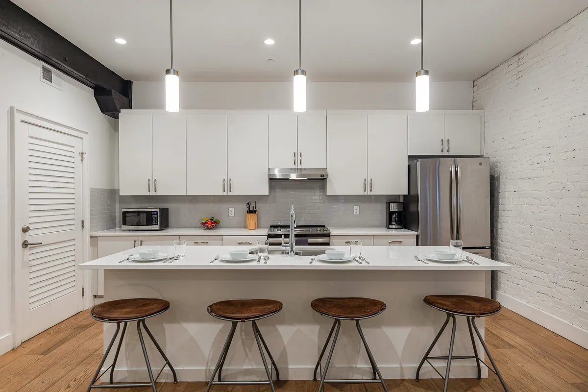 A contemporary kitchen with a central island surrounded by bar stools, white cabinets, stainless steel appliances, and an exposed brick wall. The space feels modern, warm, and perfect for entertaining or casual meals.