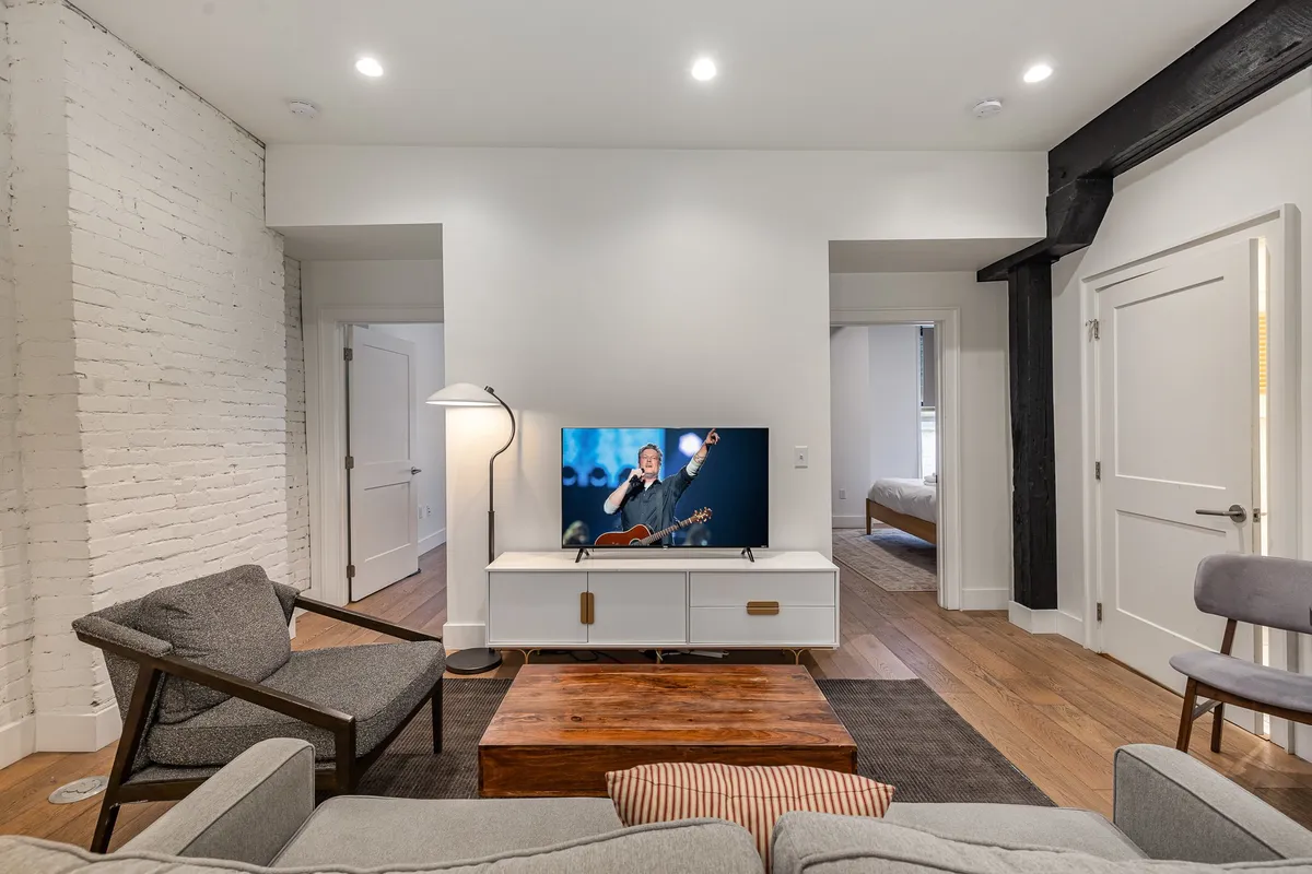 A cozy and modern living room featuring a plush sofa, coffee table, TV displaying a musical performance, and a floor lamp. The space is open and inviting, with modern decor touches and flexible seating options.