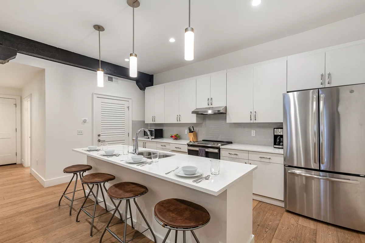 A spacious and inviting kitchen featuring an island countertop with four stools, stainless steel appliances, white cabinetry, and stylish pendant lighting. The setup is ideal for social gatherings, meal prep, and casual dining.