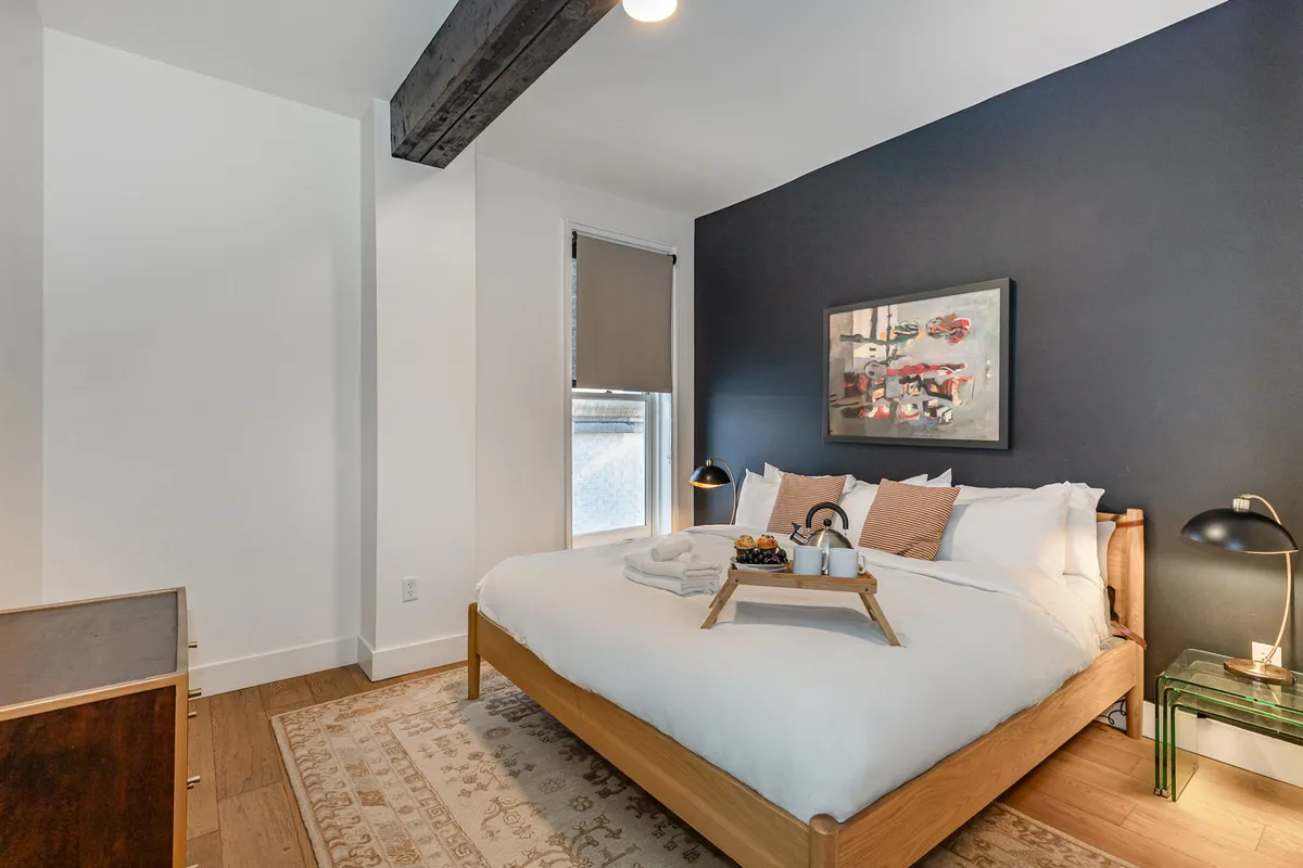 A serene bedroom featuring a comfortable bed with crisp white bedding, contemporary artwork above the headboard, soft ambient light from bedside lamps, and warm wooden flooring. The uncluttered design and inviting textures suggest a restful retreat after a lively evening.