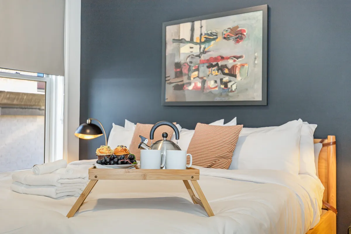 A cozy bedroom scene showcasing a plush bed with white linens, vibrant wall art, a table lamp casting a warm glow, and a breakfast tray with fresh food and drink. The setting evokes comfort and the pleasure of waking up refreshed.