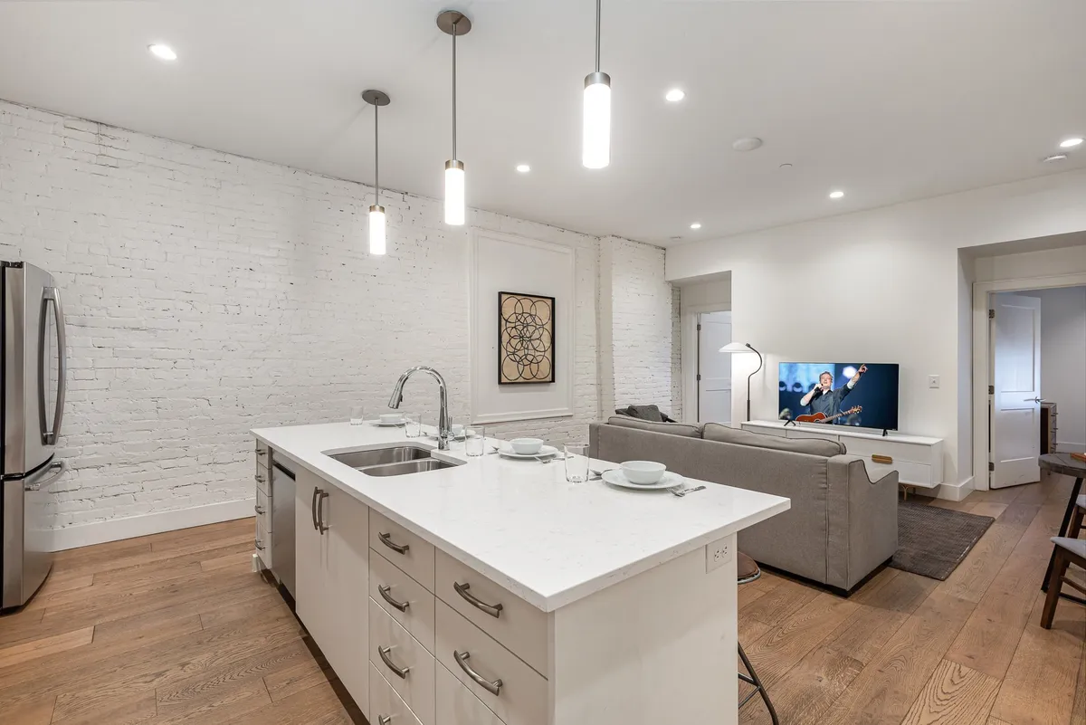 A modern open-concept living area featuring a kitchen island, sofa, television, and an exposed brick wall. The layout encourages interaction and multitasking from cooking to watching TV, with ample space for group activities.