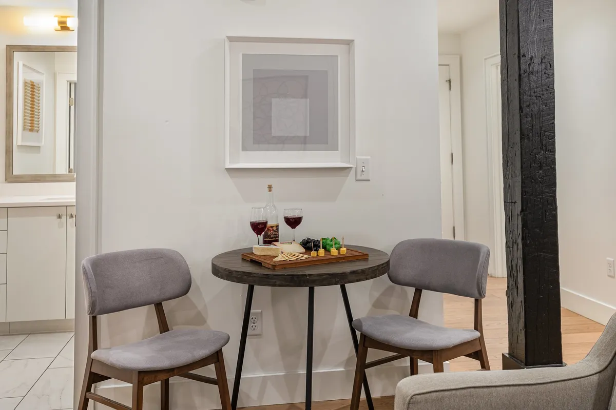 A cozy dining nook with a small round table, two wine glasses, and a snack platter, framed by artwork and a rustic wooden beam. The setting feels intimate and ideal for quiet conversation or celebratory toasts.