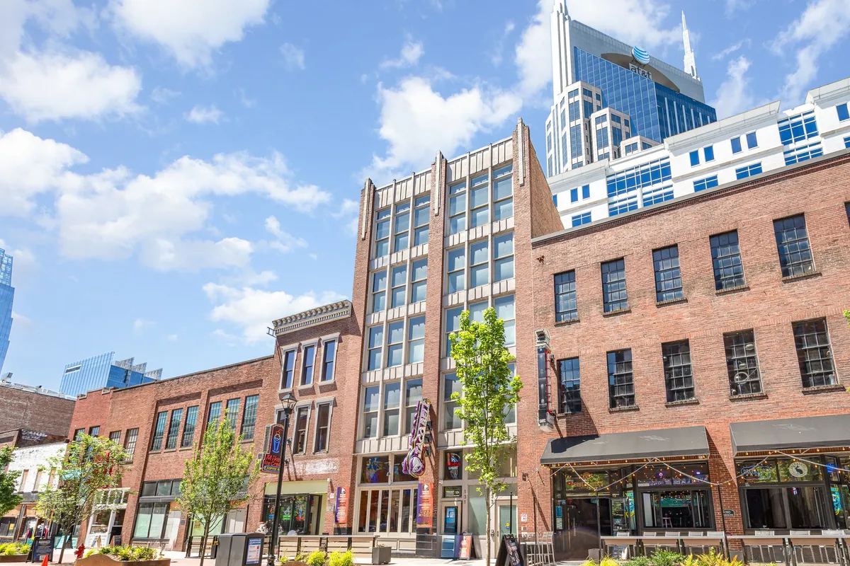 This image captures a lively street scene featuring historic brick buildings alongside modern skyscrapers, with green trees and an outdoor seating area. People are shown shopping, dining, and socializing, creating a dynamic urban backdrop.